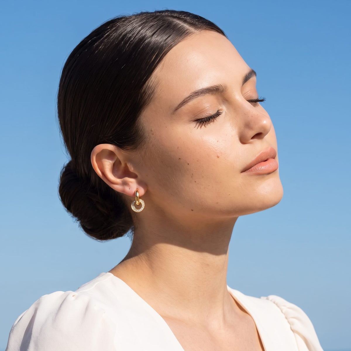 Woman wearing the Clémence circle huggie gold earrings on the ear against a bright blue sky