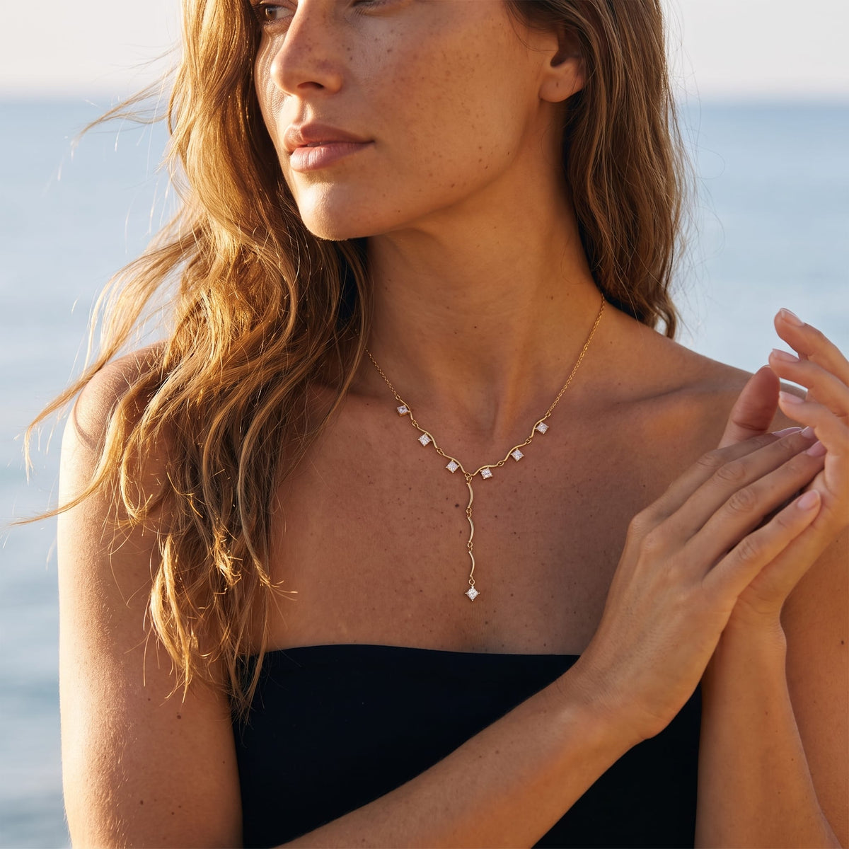 Woman wearing the Gemma square crystal gold necklace at the collarbone with the blue ocean behind her