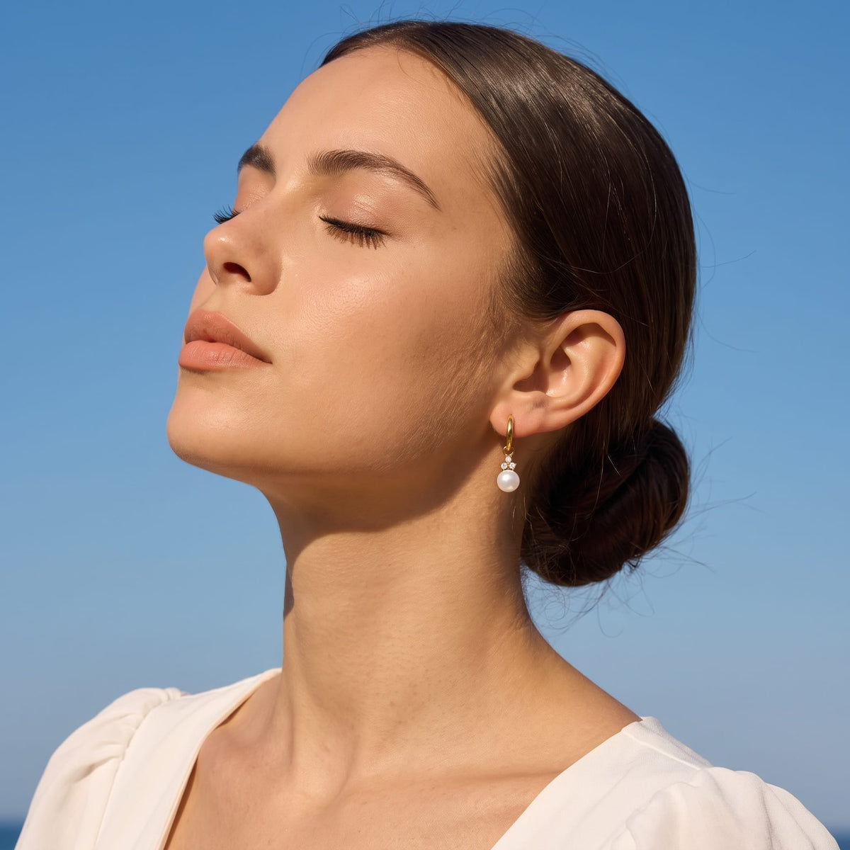 Woman wearing the Hortense pearl huggie gold earrings on her ear under a blue sky