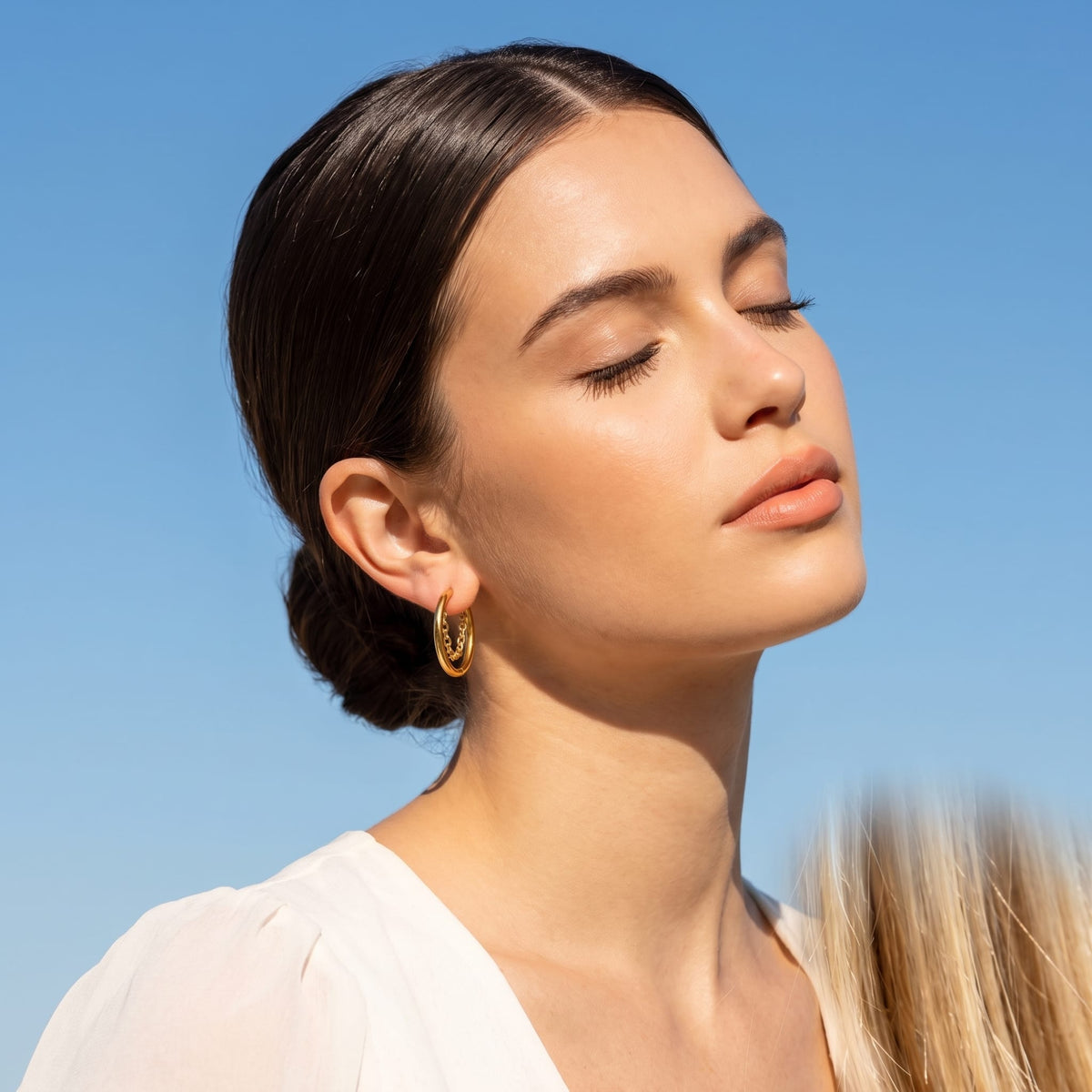 Woman wearing the Romy chain hoop gold earrings on the ear, tilted upward against a clear blue sky