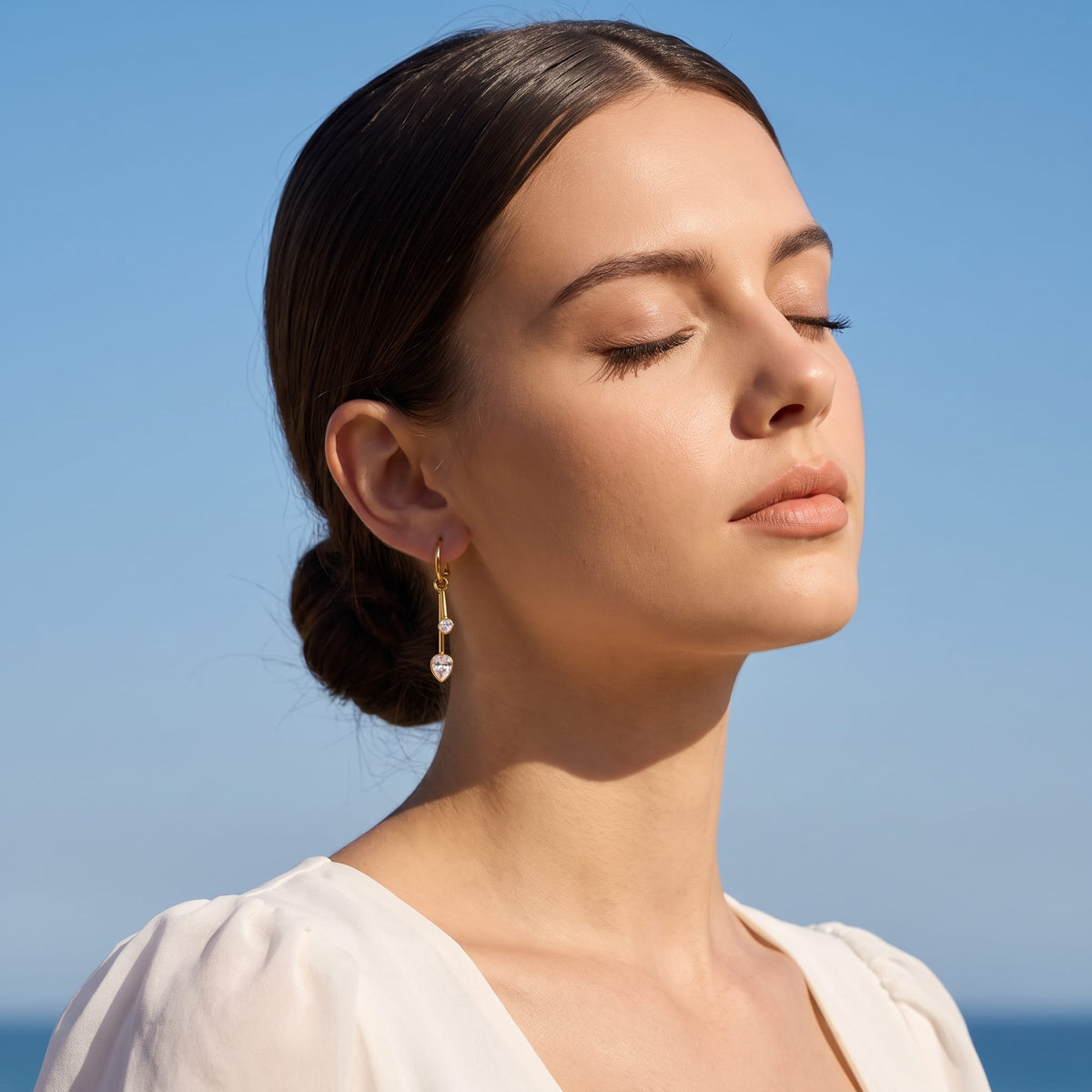 Woman wearing the Sixtine crystal bar gold earrings on her ear with eyes closed in sunlight