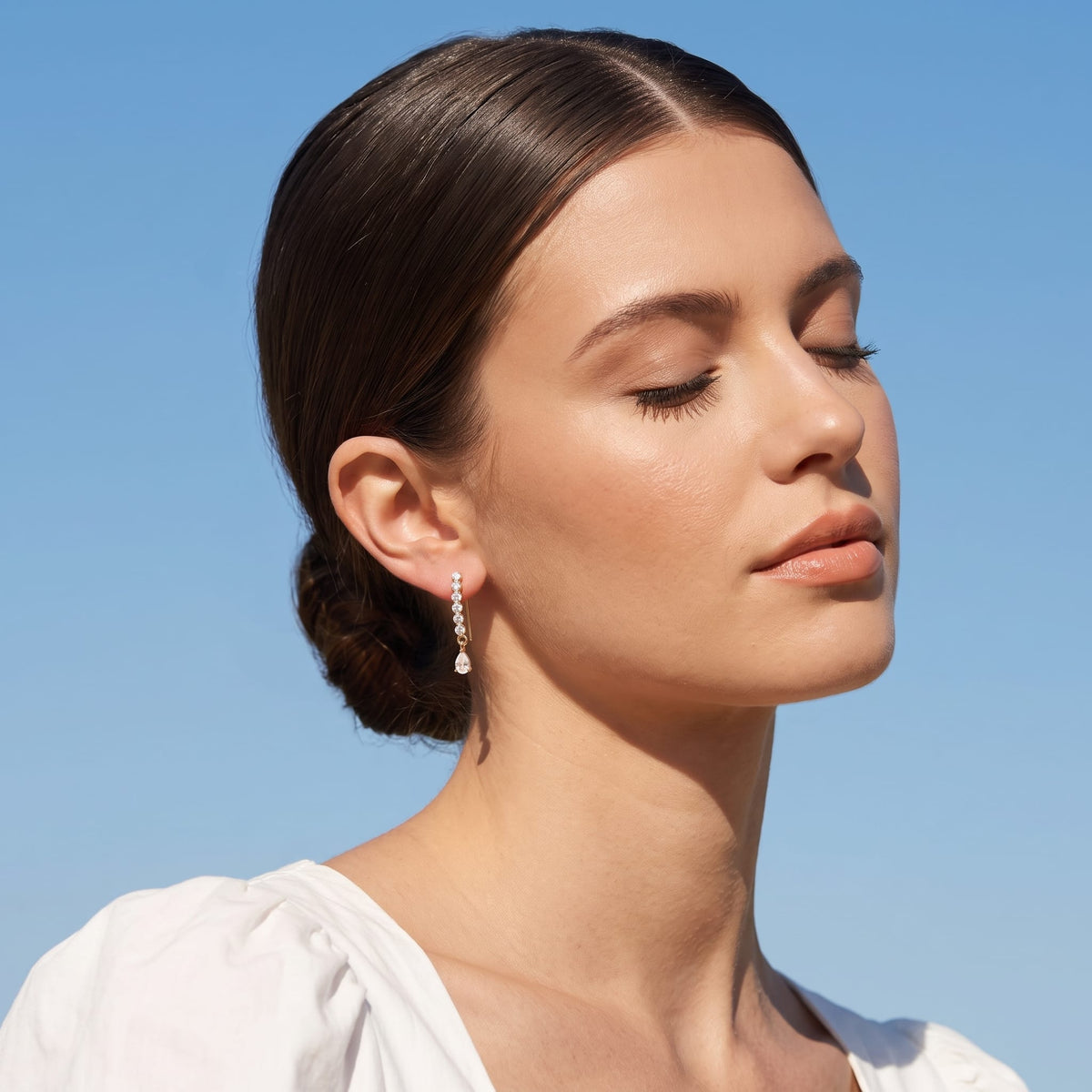 Woman wearing the Sol(ne crystal drop gold earrings on the ear, facing right with closed eyes against a blue sky