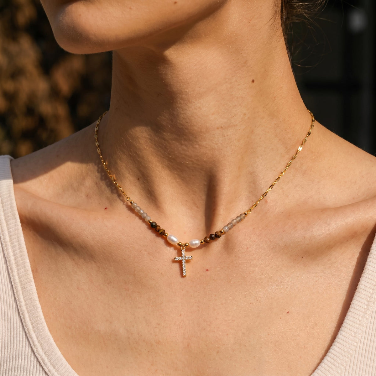 Woman wearing the Viola pearl cross gold necklace at the collarbone in warm sunlight