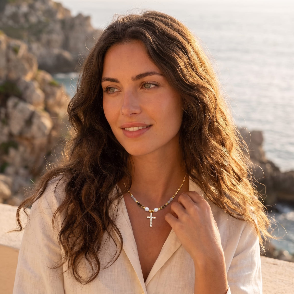 Woman wearing the Viola pearl cross gold necklace at the collarbone outdoors near the sea