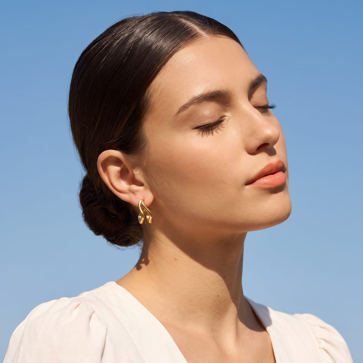 Woman wearing the Anouk double wave gold earrings on the ear against a bright blue sky background