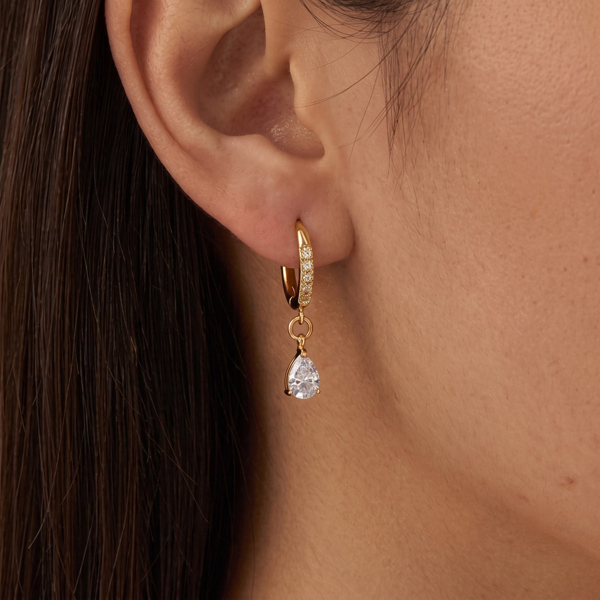 Dark-haired woman wearing an Aurélie gold huggie with a clear teardrop crystal swinging below