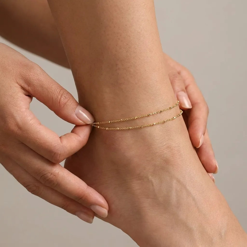 Hands adjusting the Wren layered chain anklet, two fine gold beaded chains resting on the ankle