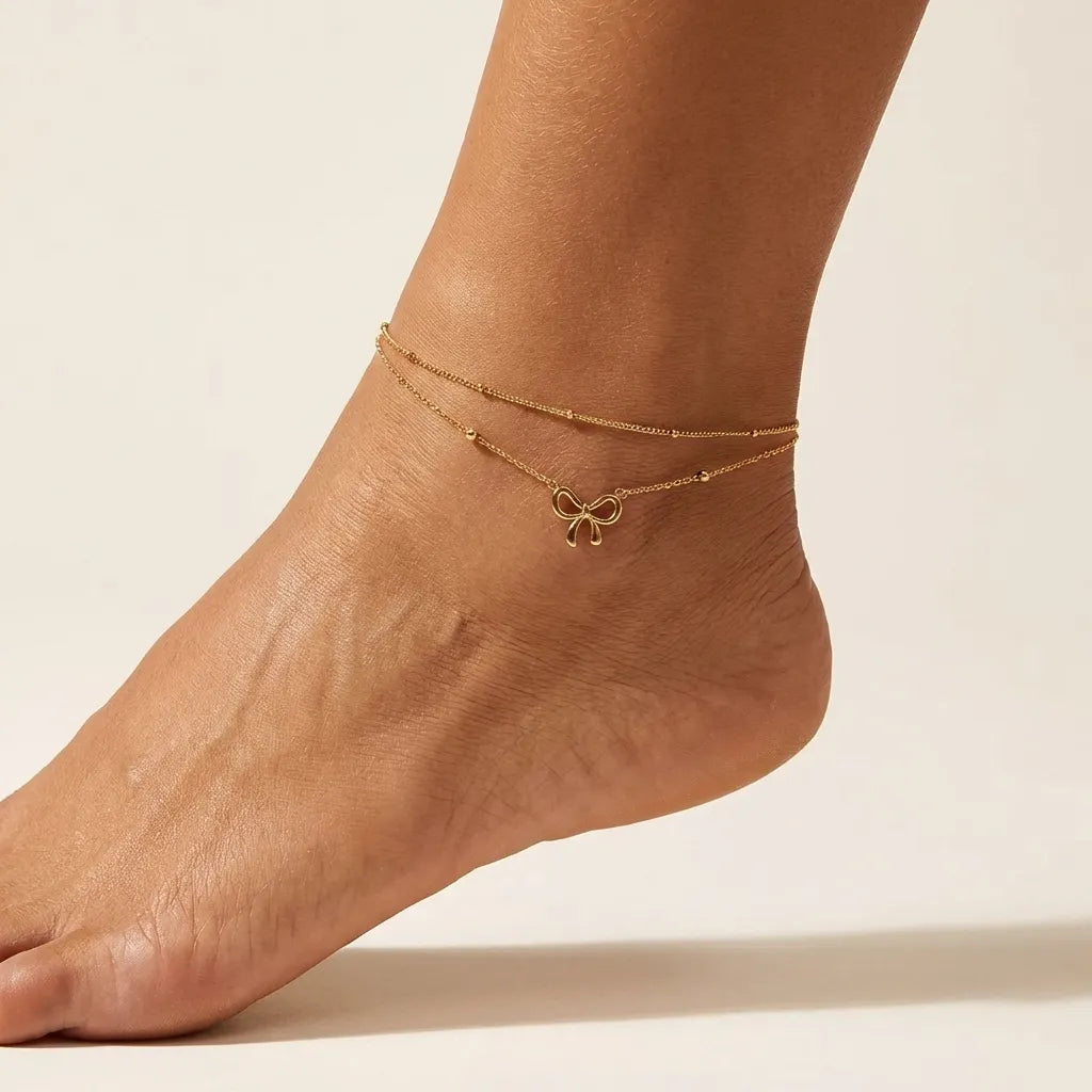 Woman wearing the Aurélie gold bow anklet, two beaded chains with an open bow charm on the ankle