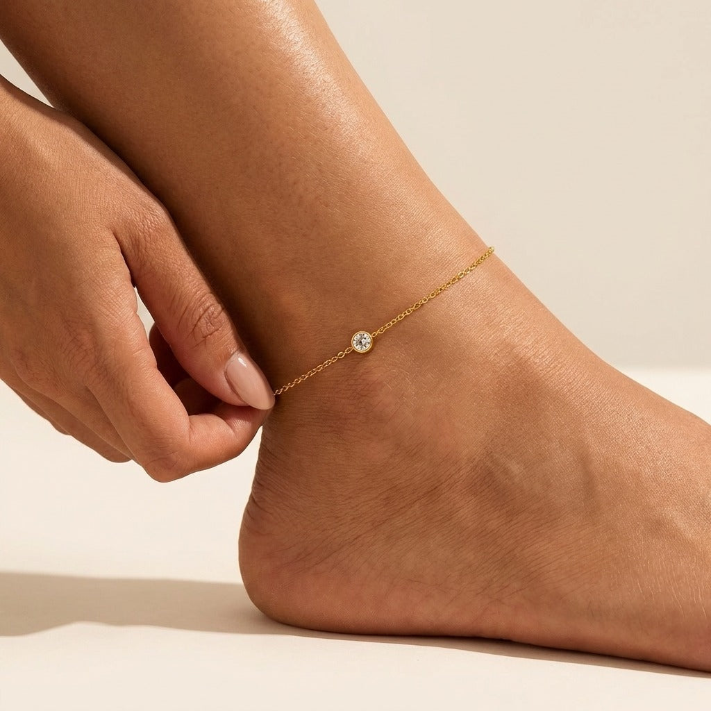 Woman adjusting the Éloise crystal anklet in gold, a single bezel-set crystal on a fine chain