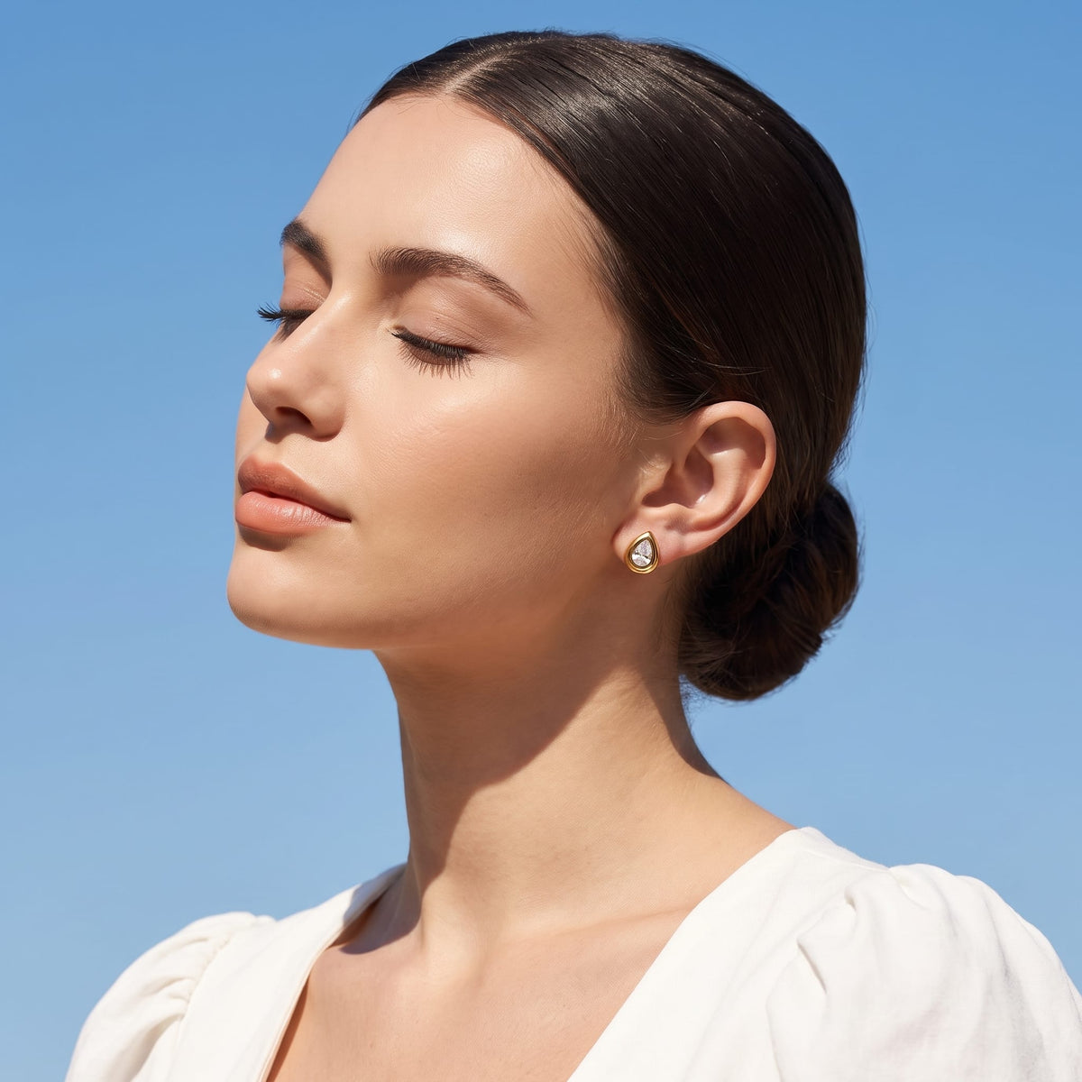Woman wearing the Élodie teardrop bezel gold earrings on the ear against a clear blue sky