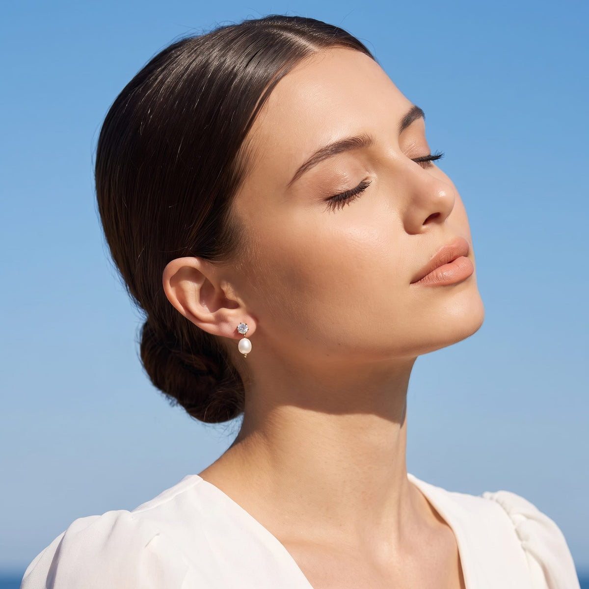 Woman wearing the Ginevra pearl drop gold earrings on the ear with eyes closed in a white dress