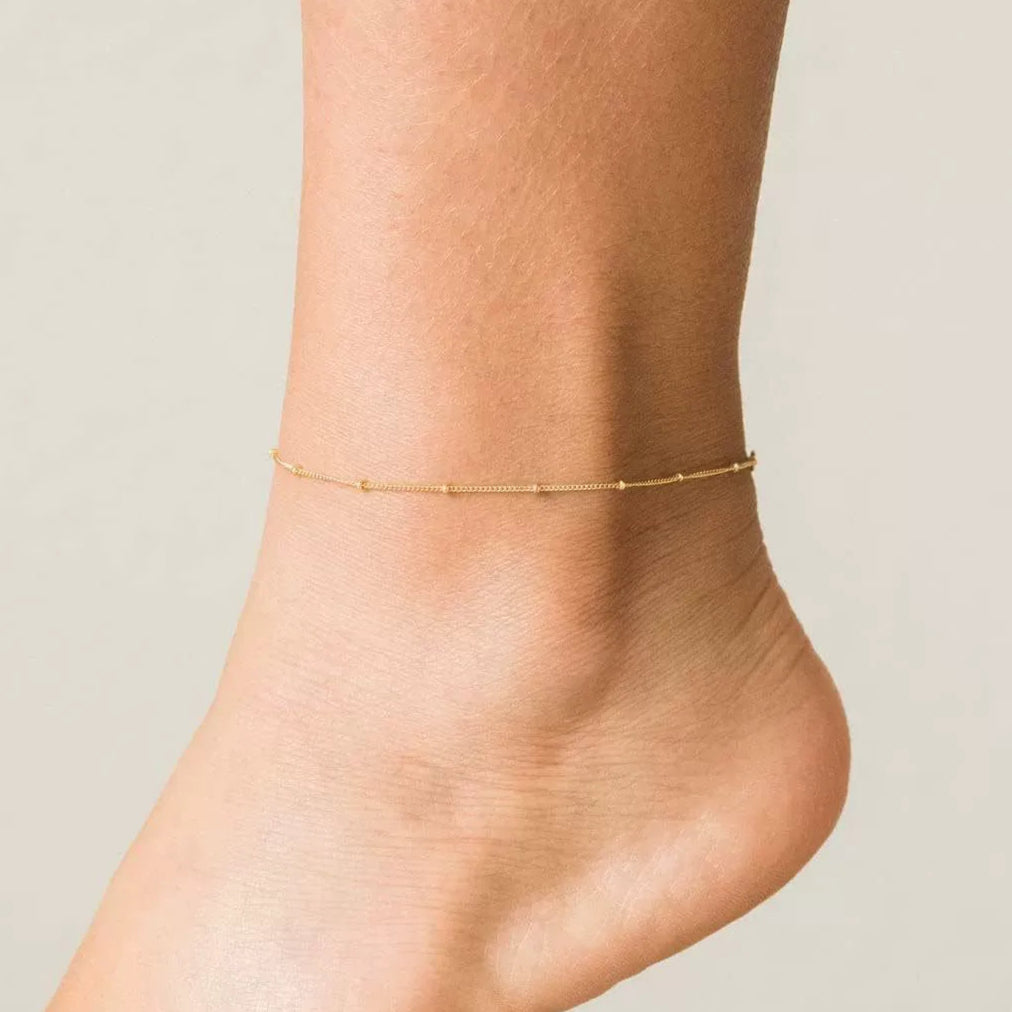 Woman wearing the Wren gold anklet, a fine satellite chain with tiny beads, just above the heel
