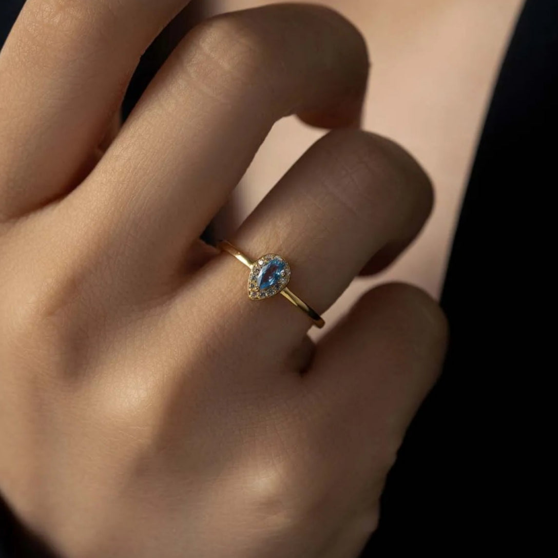 Hand wearing the Isabella radiant crystal ring in gold with a blue pear stone framed in crystals