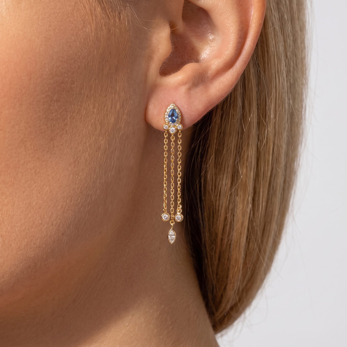 Woman wearing an Isabella gold drop earring with a blue pear stone and fine crystal-tipped chains