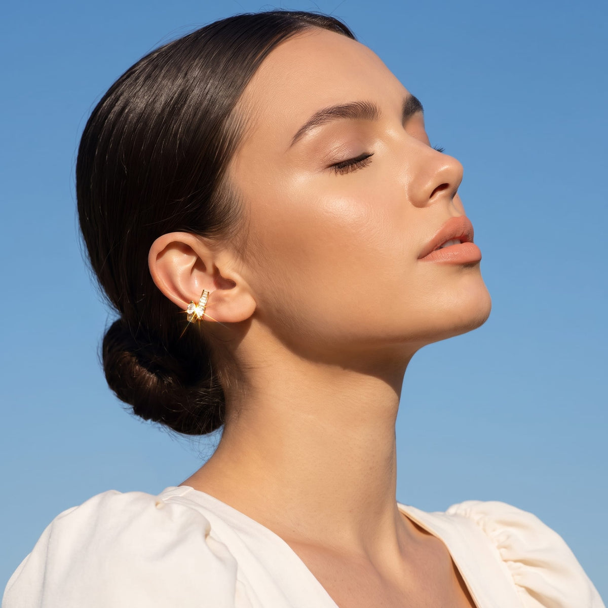 Woman wearing the Laure baguette gold ear cuffs on her ear under a blue sky