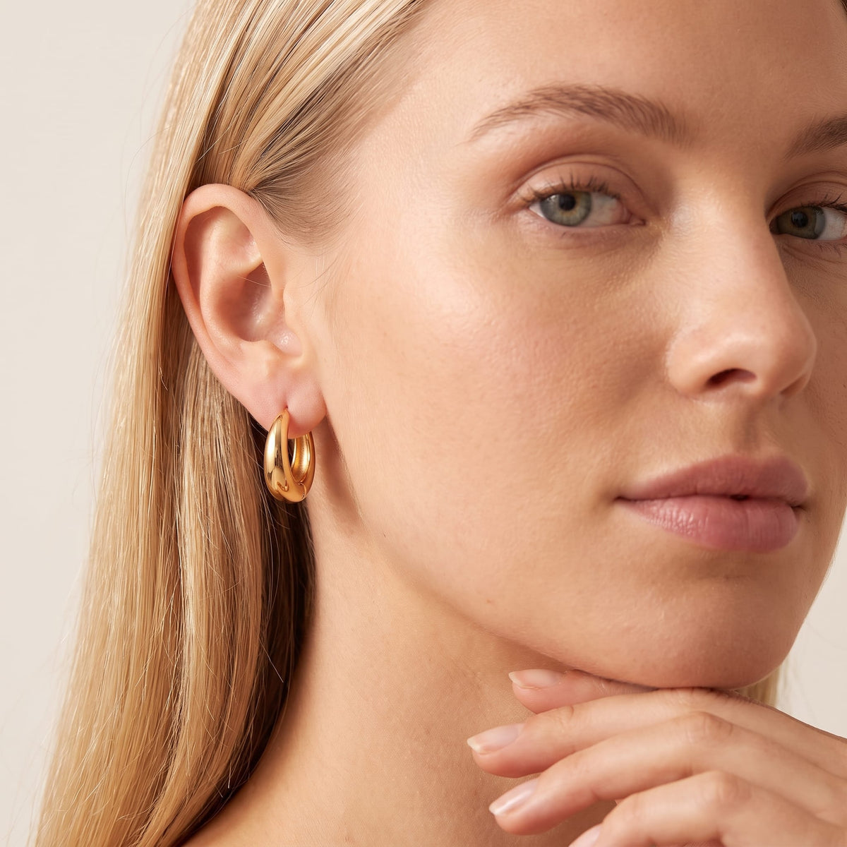 Woman wearing the Nina dome huggie gold earrings on the ear with hand resting under her chin