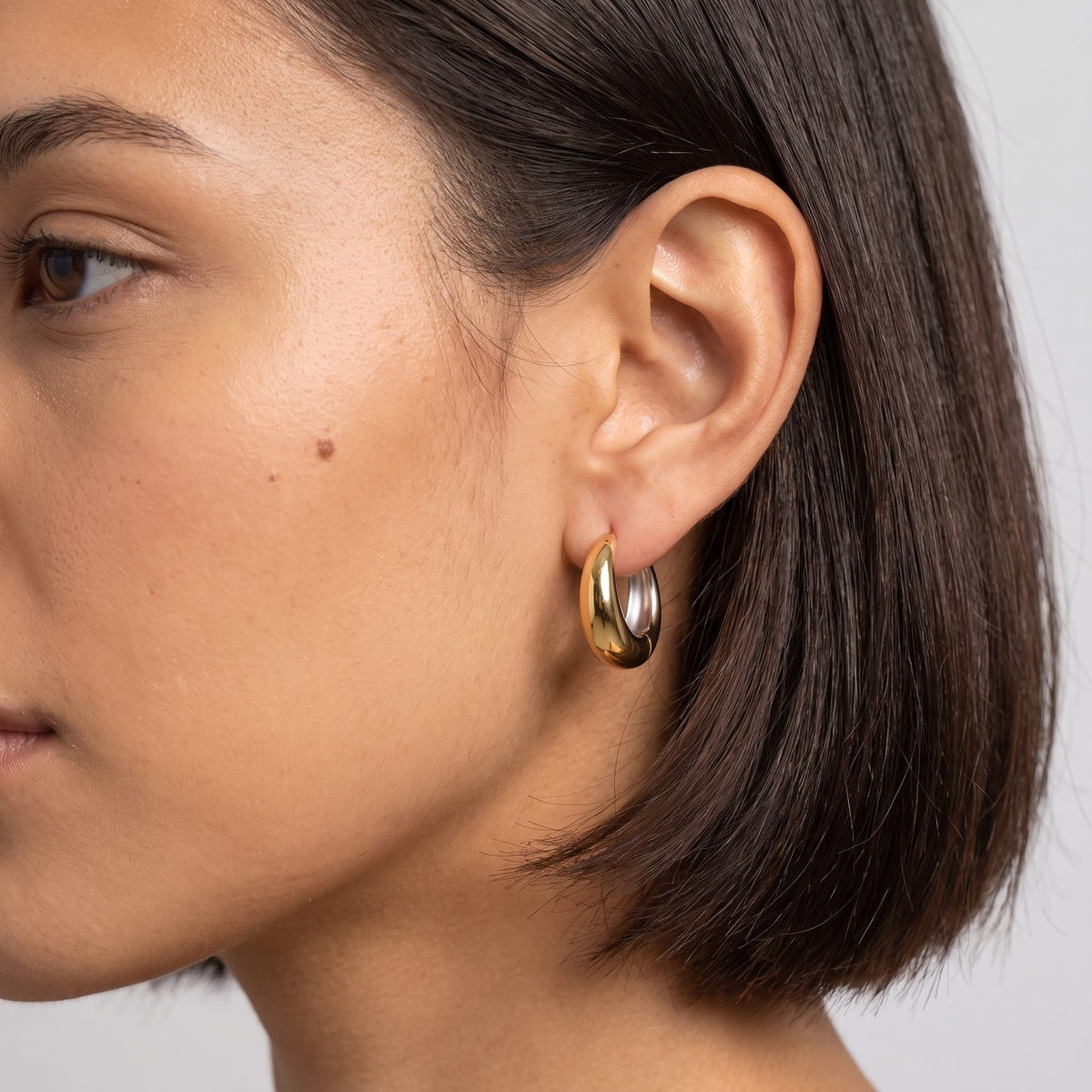 Woman wearing the Nina dome huggie two-tone earrings on the ear in a close up studio shot