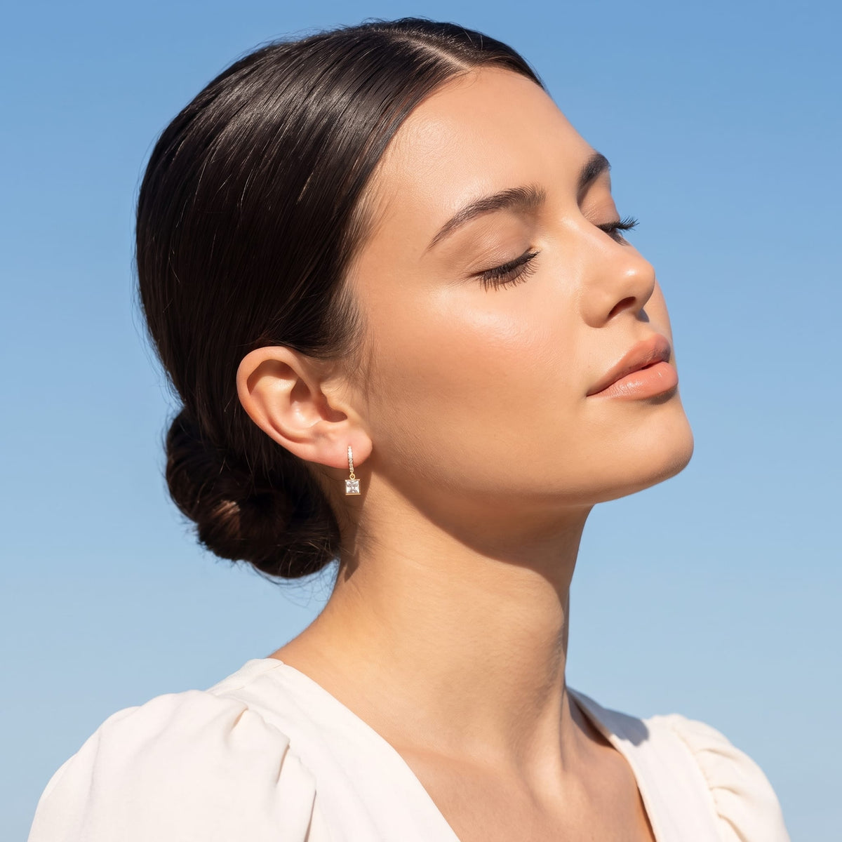 Woman wearing the Paola crystal square gold earrings on the ear under a blue sky