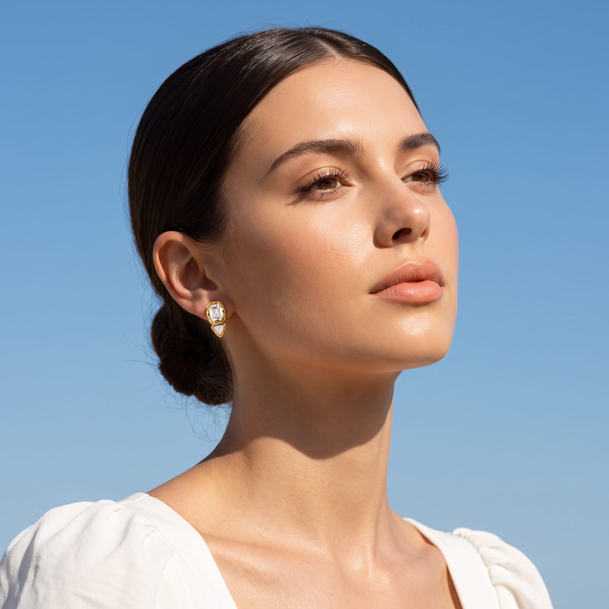 Woman wearing the Sidonie emerald-cut crystal gold earrings on her earlobe against a blue sky