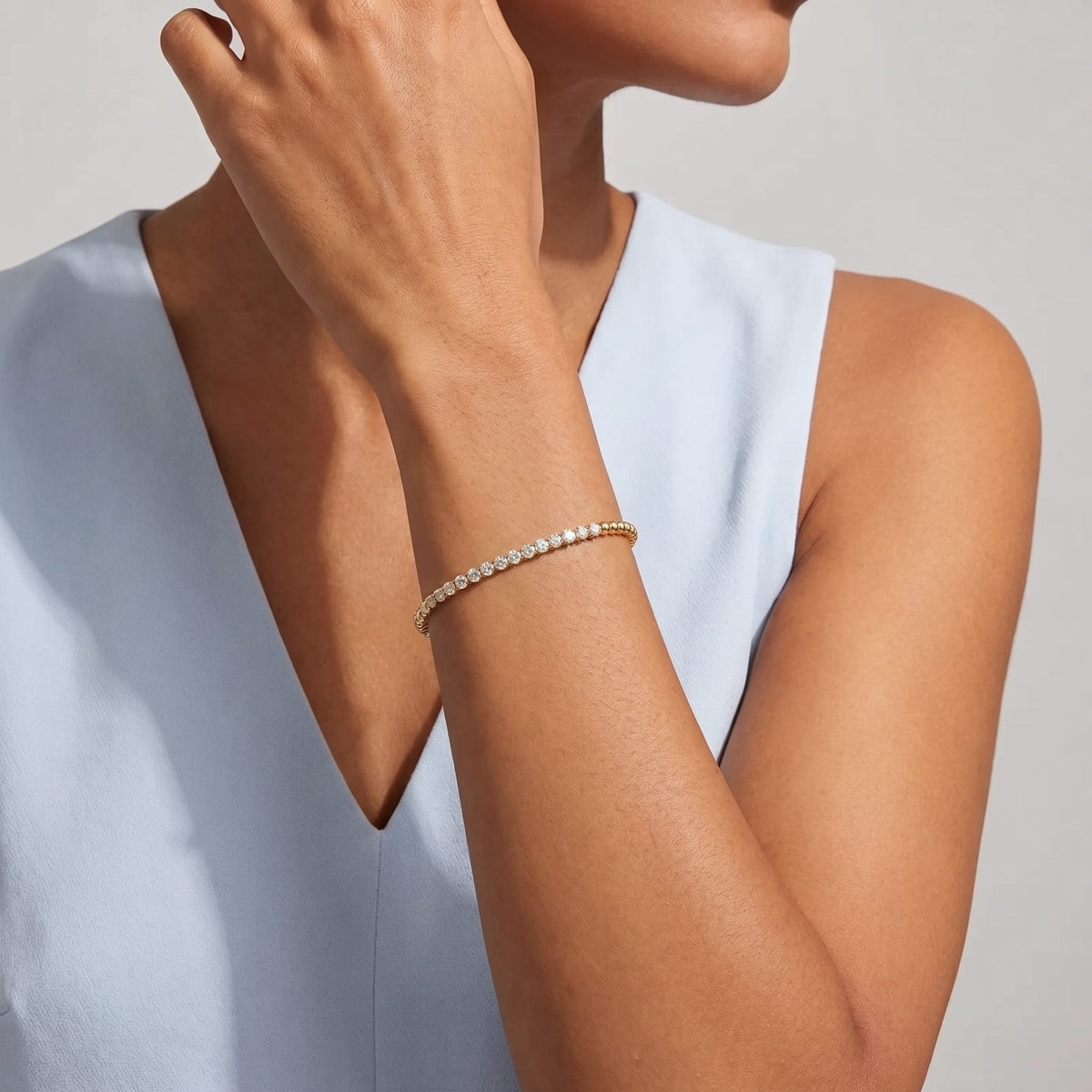 Woman wearing the Vida tennis bead gold bracelet on the wrist in a blue top posing near chin